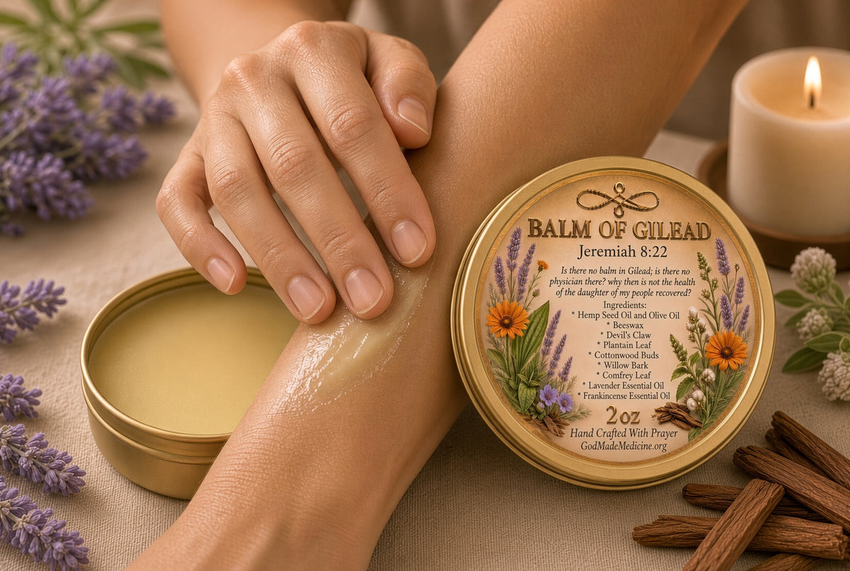 A person applies a pale yellow balm cream to their forearm while holding a round tin labeled 'Balm of Cilead'. The scene is styled with lavender flowers, cinnamon sticks, a lit candle, and a neutral fabric background