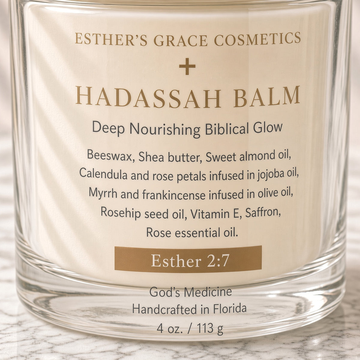 A close-up of a clear glass jar candle labeled 'Esther's Grace Cosmetics + Hadassah Balm' with a cream-colored label and gold and black text, set on a light marble surface.

