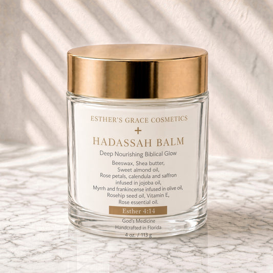 A glass jar of Esther's Grace Cosmetics Hadassah Balm with a metallic gold lid and white label, set on a light marble surface with soft leaf shadows.

